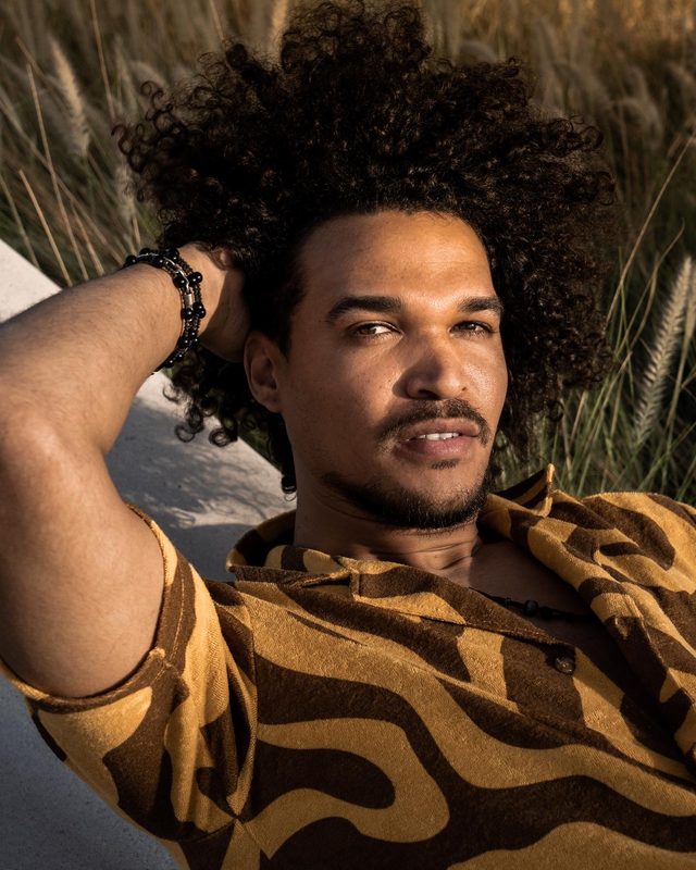 Gabriel Oramas lying back in tall grass with one arm behind his head, looking at the camera