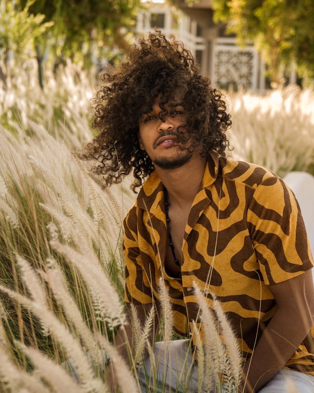 Gabriel Oramas sitting among pale feathery grass, hair partly over his face, looking at the camera
