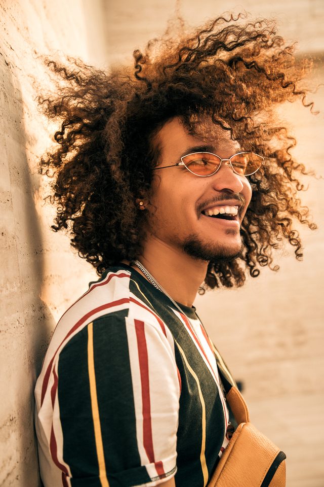 Gabriel Oramas laughing against a wall, wearing orange-tinted sunglasses and a striped T-shirt